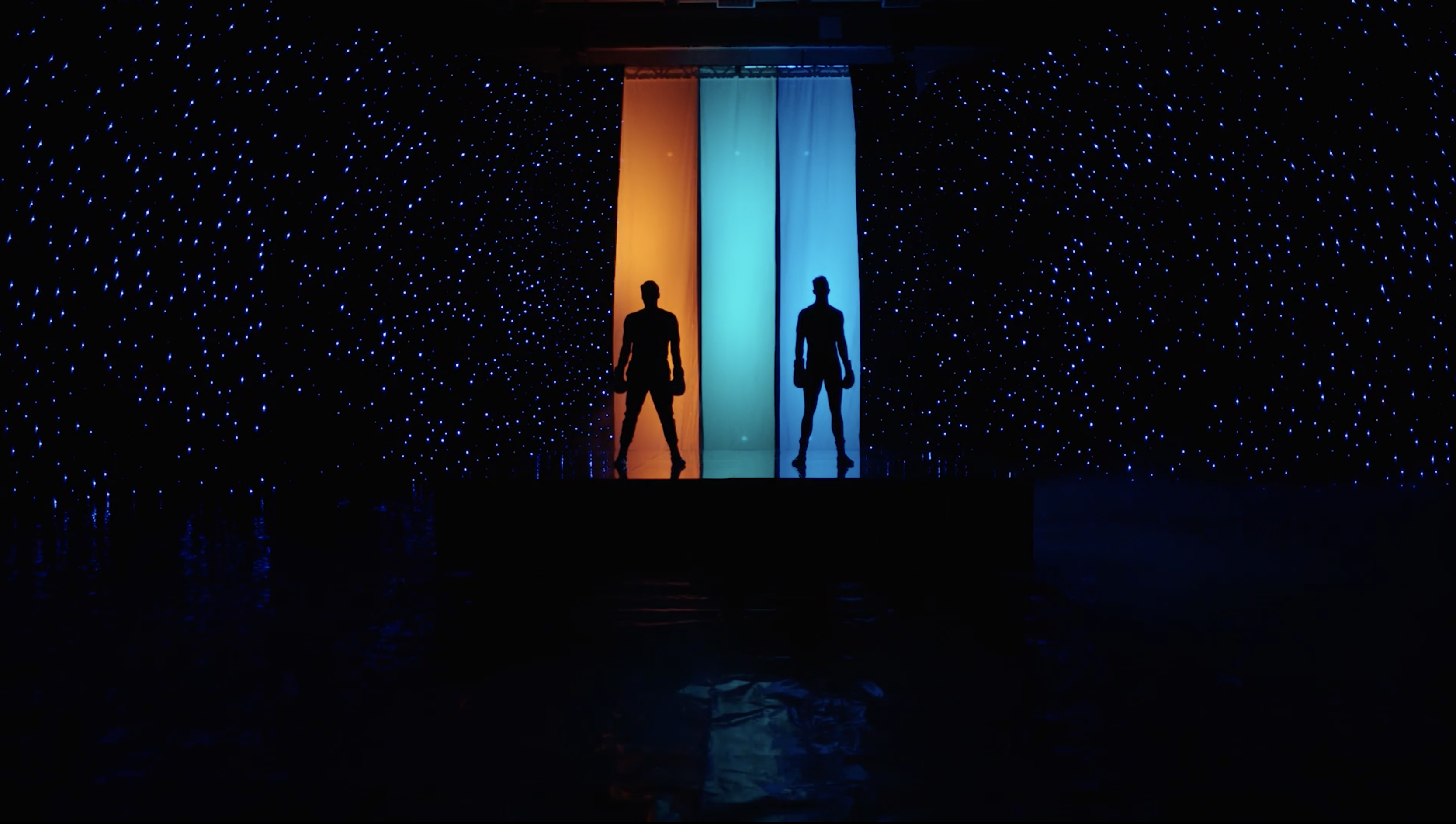 Two men silhouetted in front of orange and blue draped banners